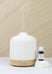 White textured diffuser with wooden base on a light surface, next to a small bottle of essential oil.