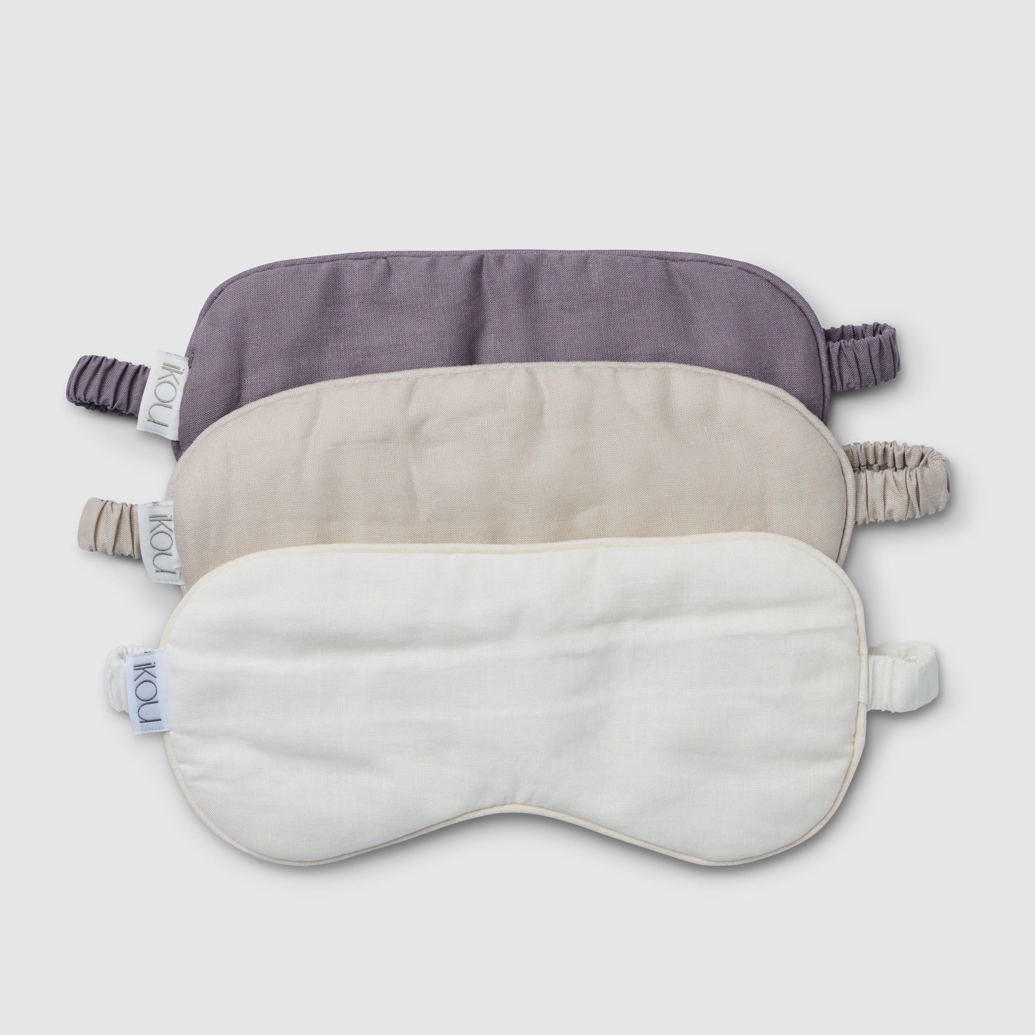 Set of three eye masks in purple, beige, and white on a light gray background